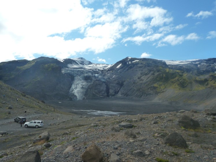 A big hole in the landscape between a glacier-topped mountain and a grey riverbed. There are a couple of vehicles parked on the edge showing just how big this hole is. It was once a lake but it's now filled up with volcanic ash.