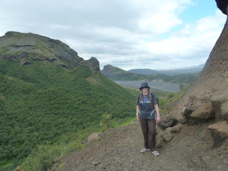 Me, in sandals, standing on the top of a mountain with a great green-covered mountain to my left and the grey riverbed behind me.