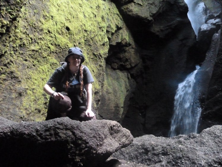 Me crouched on a boulder in a cave with green-coated walls and a waterfall coming down the right-hand side.