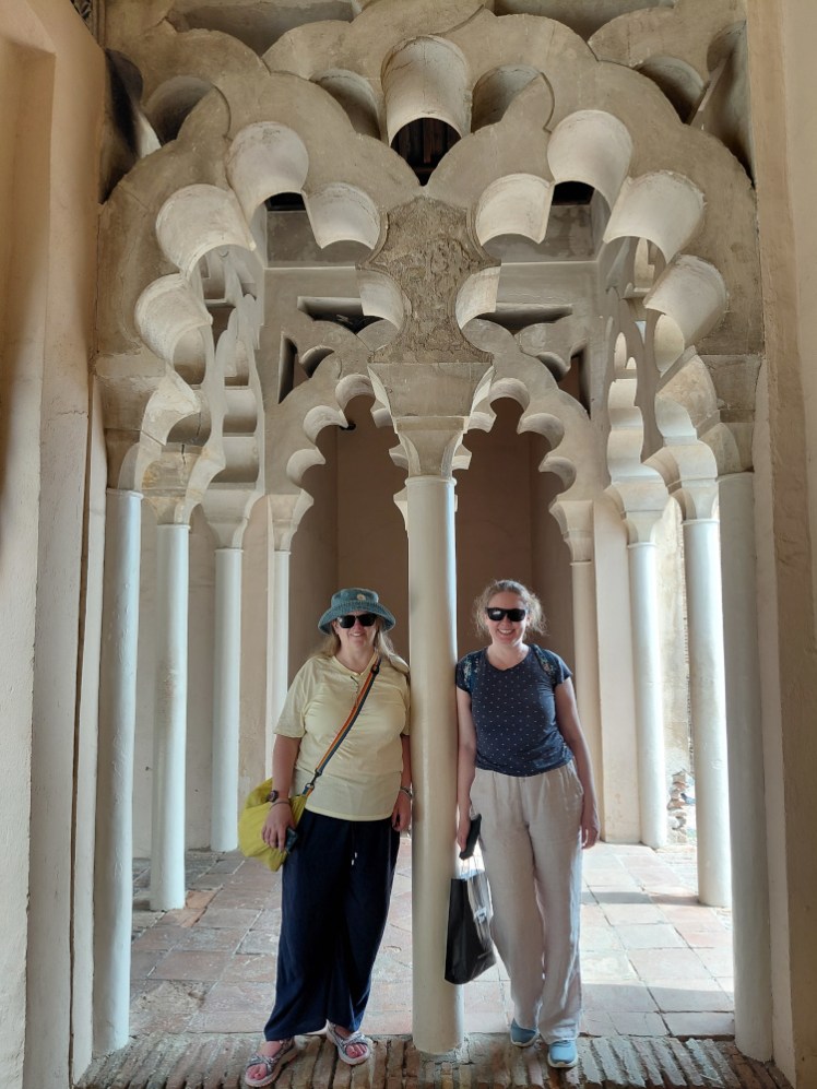 Me in the pink sandals at Malaga's Alcazaba with Esther - she's in light trousers and a darker top, I'm in darker trousers and lighter top and we're leaning against a pillar under decorative arches, looking very symmetrical.