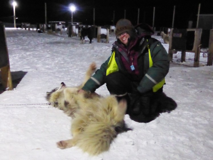 Me in a padded snowsuit kneeling on the floor playing with a very fluffy sled dog lying with his belly up for cuddles.
