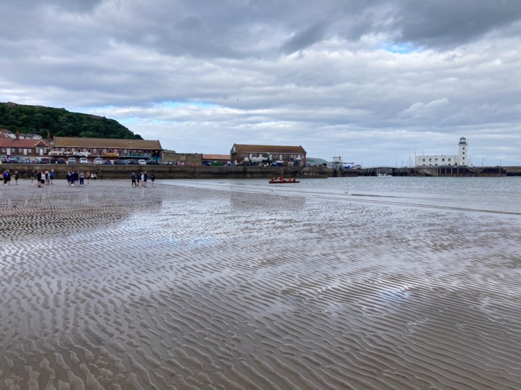The beach at Scarborough at low tide. The retreating water has scoured the sand into ridges and some amateur rowing club boats are coming ashore to a welcome of fans.
