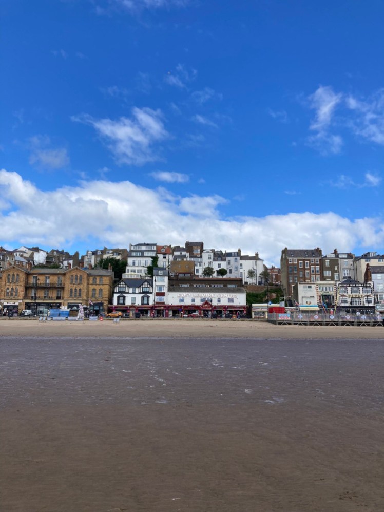 The seafront at Scarborough, with blue sky above and sand below and a big arcade in the row of shops in the middle.