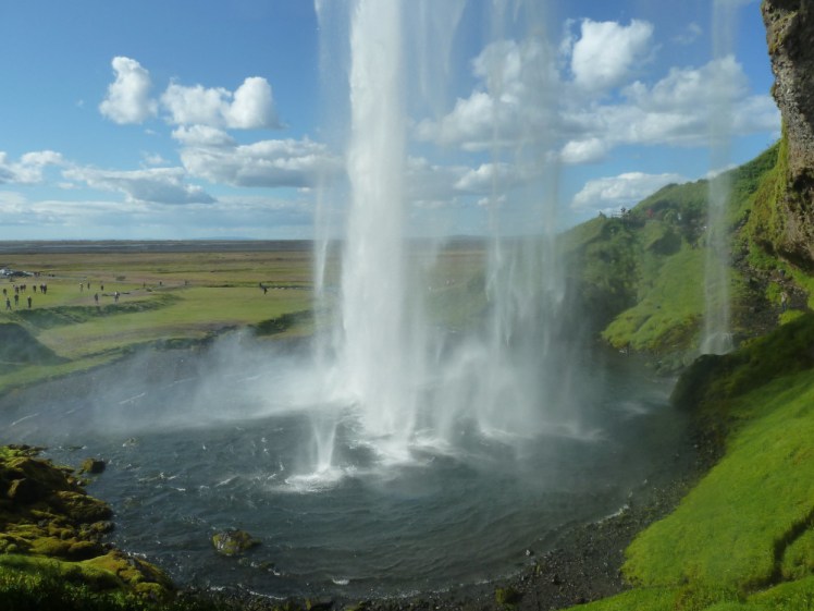 A view from behind Seljalandsfoss, the water falling into a pool and gently lit by late afternoon sunlit turning the landscape here green.