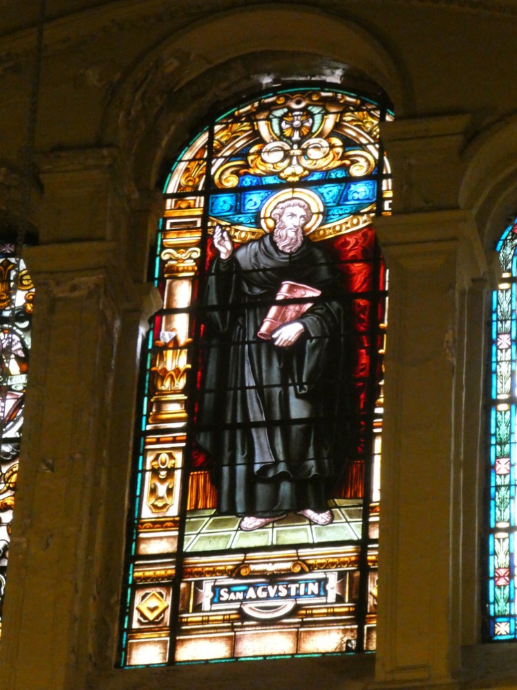 A stained glass window depicting St Augustin in black flowing robes. He's standing against a rich red background and gold pillars and arches over him.