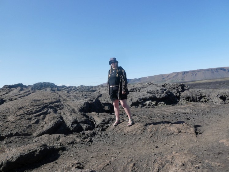 Me on a lava field under blazing sunshine and blue sky, wearing shorts, a short black t-shirt, an oversized brown checked shirt and green sandals that almost blend in with the background.