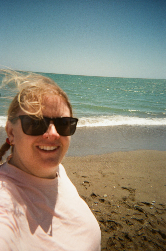 A selfie on the beach in a pale pink t-shirt with a turquoise sea behind me.