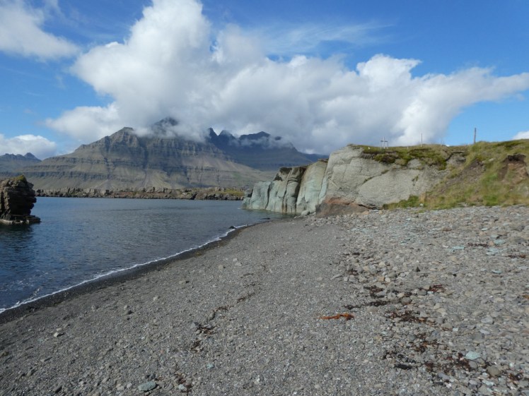 A gravelly beach that ends in a bright green cliff. Behind that is a mountain with a cloud sitting on top of it.