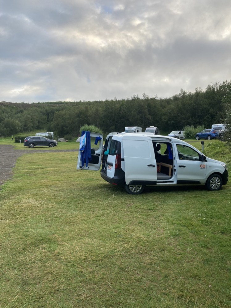 My campervan parked on the grass at Systragil with forest in the background. The doors are all open and my swimming stuff is draped over the doors to dry.