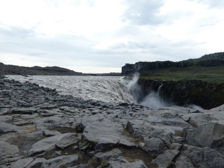 Dettifoss from the east side, a huge grey-white waterfall falling into a chasm that you can't see because of the grey rocky edge. Opposite is the green bank.