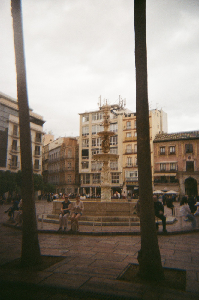 Esther & Catherine sitting on the edge of the fountain in Plaza de la Constitucion, with two palm trees framing them.