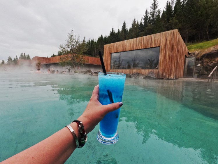 My hand in the Forest Lagoon, holding up a large cup of somehting bright blue in front of the blue-green water and the wooden sauna hut.