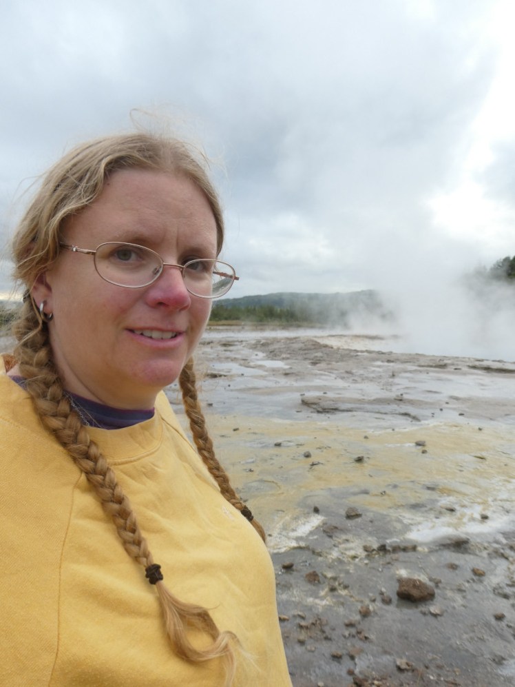 A selfie wearing my yellow jumper next to Strokkur which is gently steaming after an eruption.