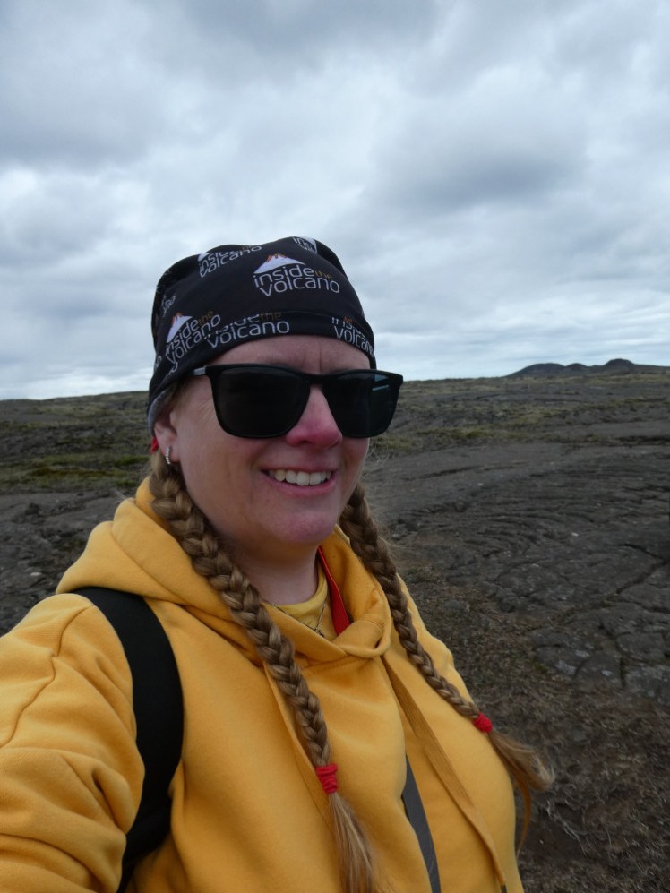 A selfie in a yellow hoodie and Inside the Volcano buff, hiking across a lava field to a volcano in the distance.