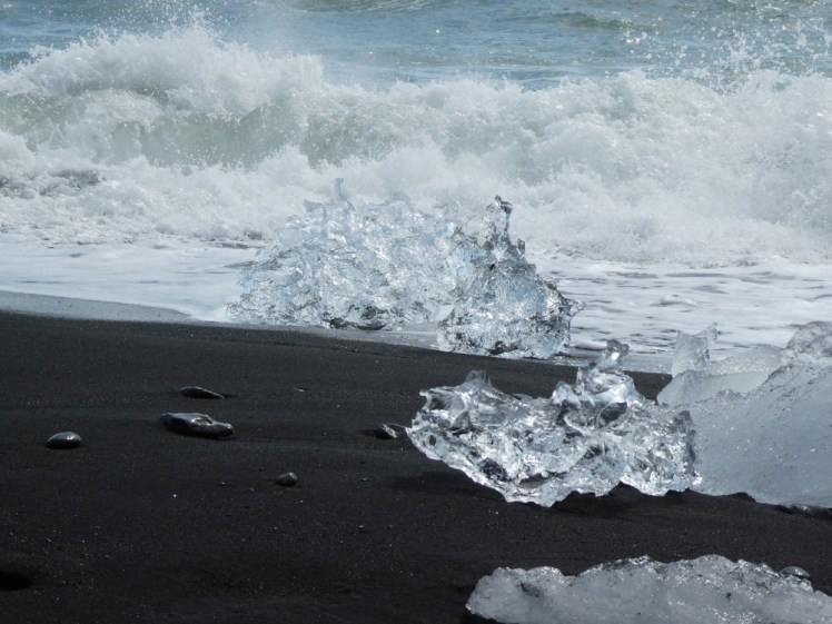 A wave breaking in the background, behind chunks of crystal-clear ice on black sand.