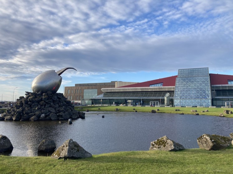 Keflavik Airport as seen from the front. A claw sticks out of a giant stainless steel egg in the middle of a pond and the airport, a low-ish building with a red roof and a glass panel along the front is visible in the background.