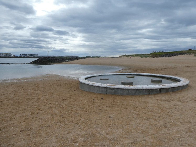 The beach hot tub up close, set on the golden beach, and with a puddle partly flowing from underneath it and partly from the fjord.