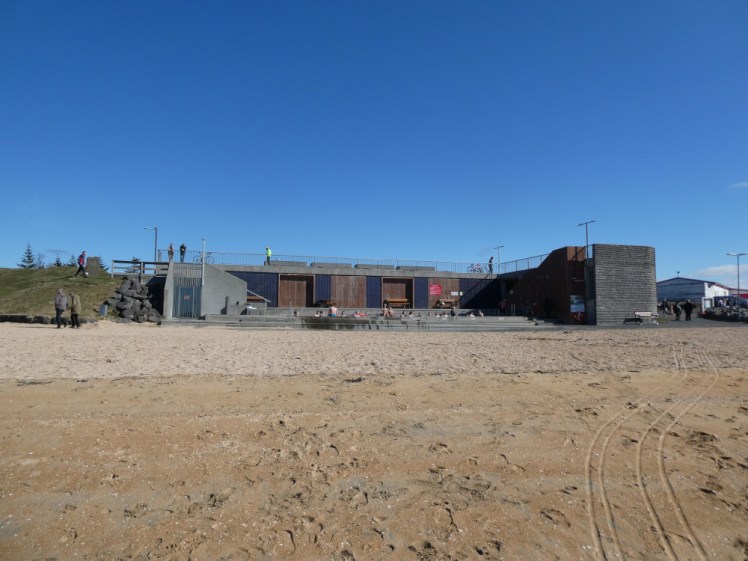 The beach club building, a single-storey building with people bathing in the long tub outside and more people enjoying the view from the roof.