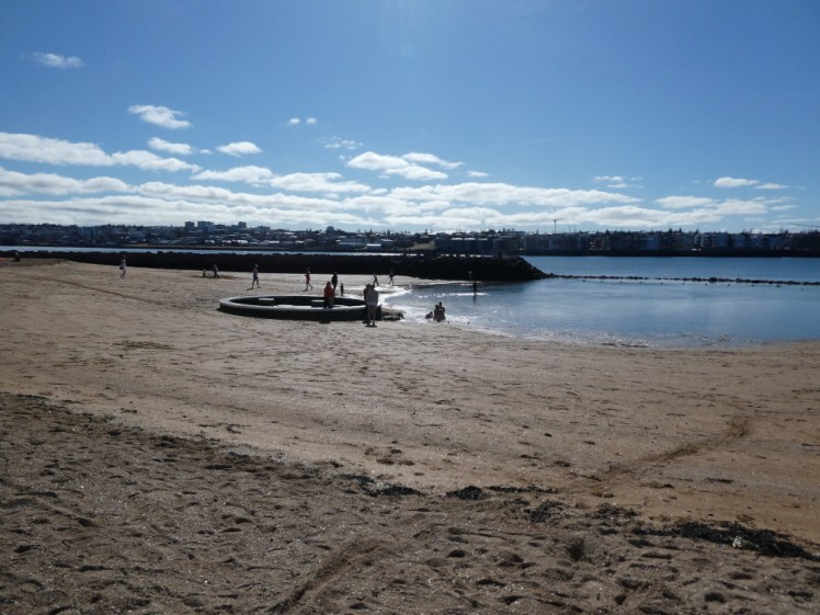 Nauthólsvík in the sun back in 2022. The sand is brownish-golden and there's a big puddle spilling over from the fjord which is separated from the beach by a low wall and a swimming lane divider.