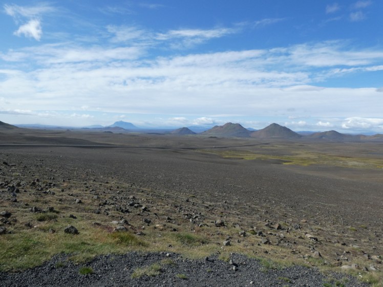 A view across the Ódáðahraun, which is just gravel and a bit of greenish lichen stretching out towards a row of small mountains.