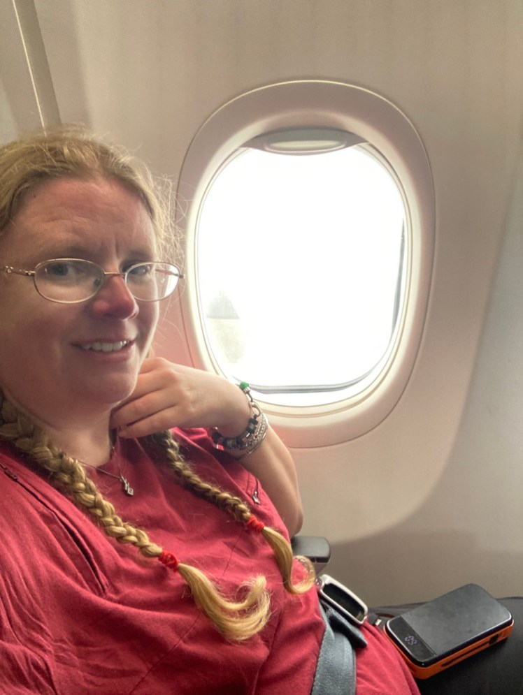 Me, now in a red t-shirt, on the plane in my window seat with my portable charger balancing on my leg waiting for the second and third buses of passengers to arrive.
