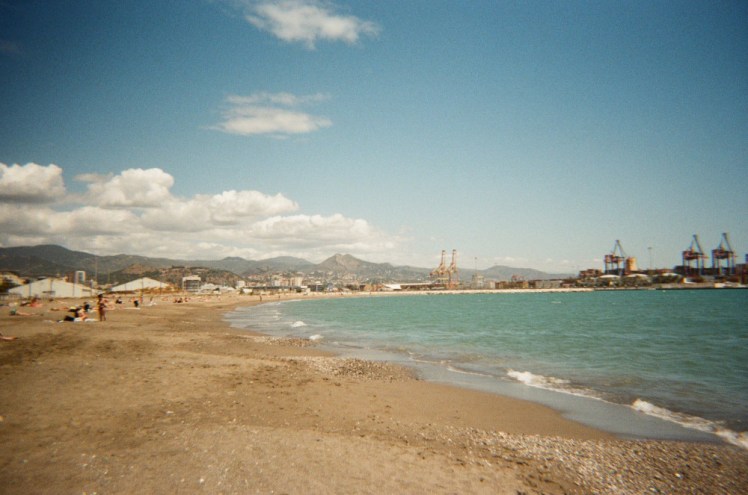 The beach, with a big blue-turquoise sea swooping up onto a golden beach with something industrial on an arm sticking out into the beautiful water.