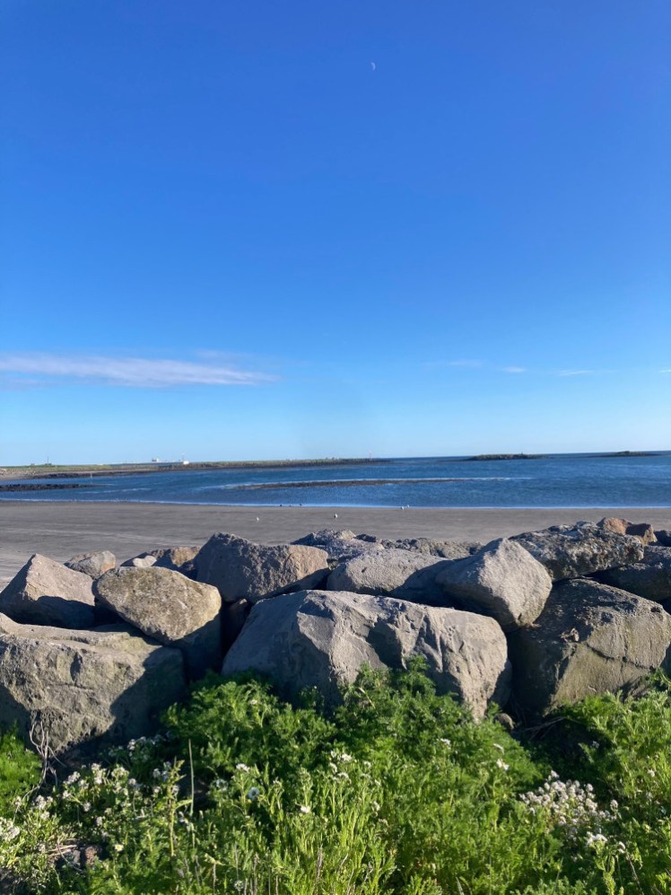 The seafront at Sandgerði, a deep blue sea and a deep grey beach under a bright blue sky.