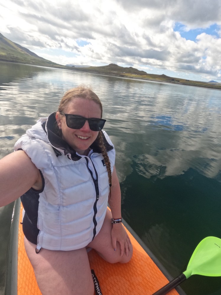 Me, in a weird buoyancy aid shaped like a giant white puffy vest, kneeing on an orange stand-up paddleboard in the fjord off the shore at Hvammsvik.