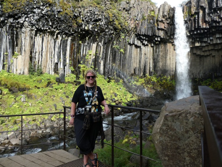 Me standing on the platform in front of Svartifoss, a waterfall falling over black basalt columns into the woods.