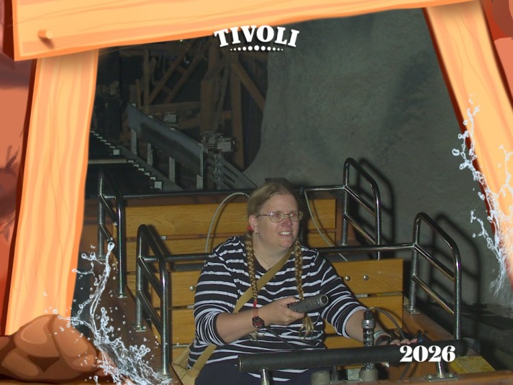 My official ride photo from The Mine. I'm sitting in a little boat with two rows of benches, pointing a laser gun shaped like a heavy metal cylinder at something the camera can't see. The picture is framed with a cartoon wooden mine support and has TIVOLI across the top and 2026 in the bottom right corner.